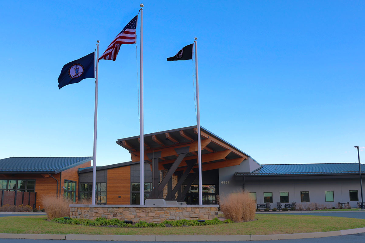 Vue extérieure du Puller Veterans Care Center, montrant l’entrée principale avec trois mâts arborant le drapeau américain, le drapeau de l’État de Virginia et le drapeau POW/MIA devant le bâtiment sous un ciel bleu clair.